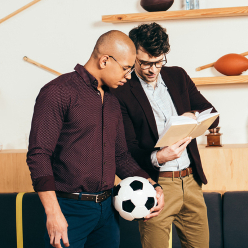 Two team managers discussing school sports team planning; one is holding a soccer ball, symbolizing athletic activities, while the other is referencing a team management guide, representing collaboration and organization.