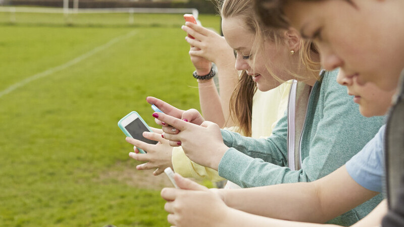 Row of five boys and girls reading smartphone text messages at football pitch